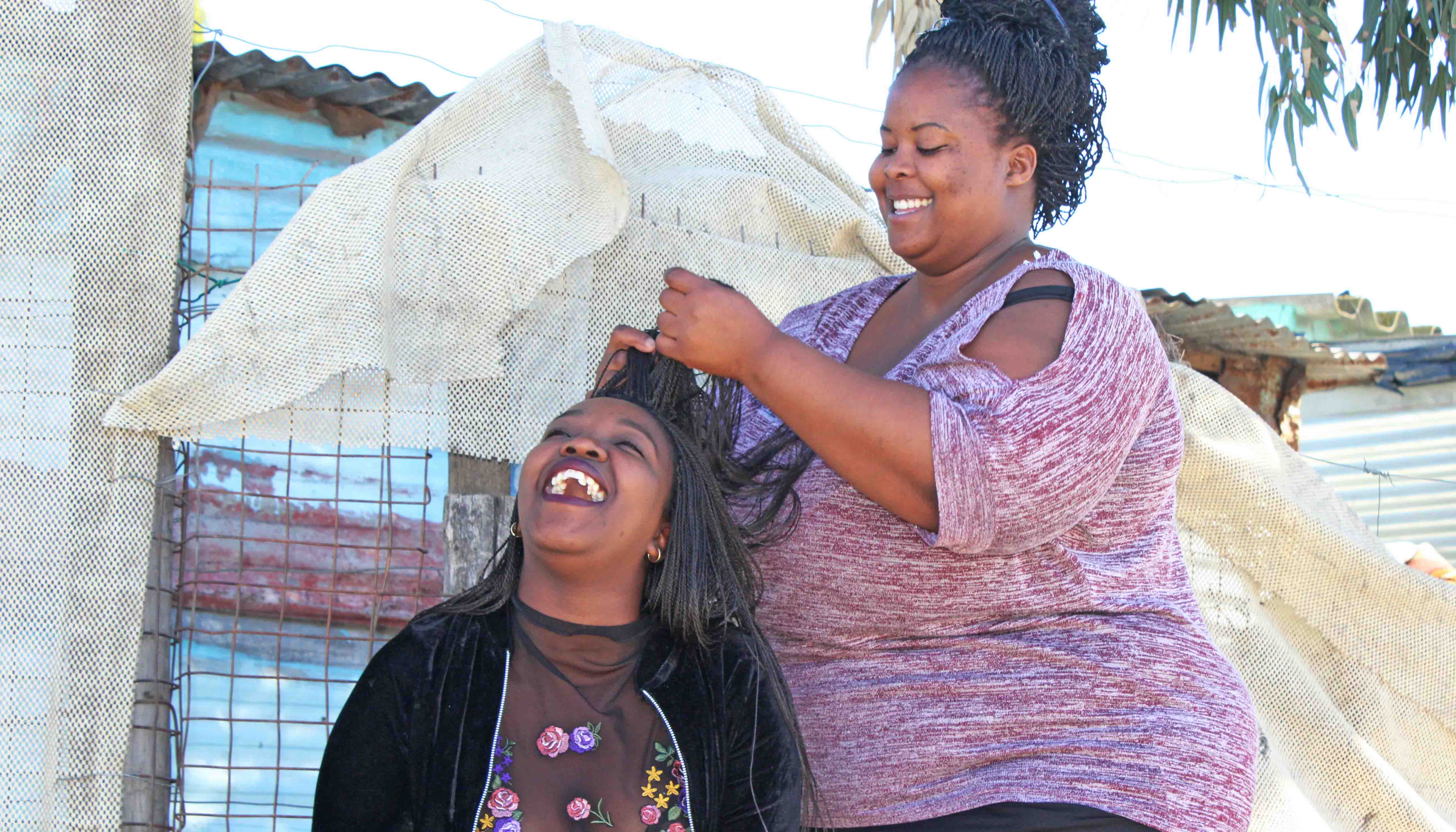 Image of a black woman smiling while braiding the hair of a younger black woman who is laughing.