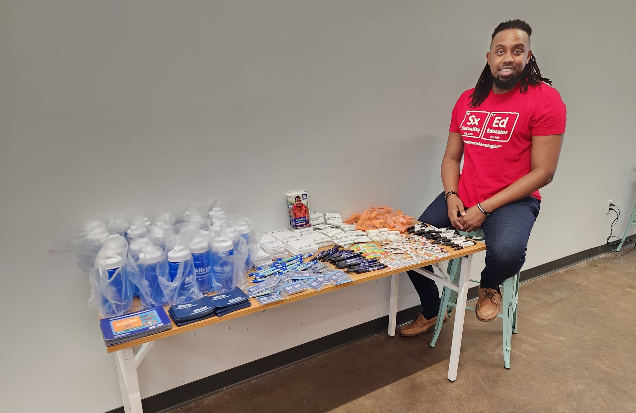 Image of JT Williams in a red shirt sitting at a table full of merch.